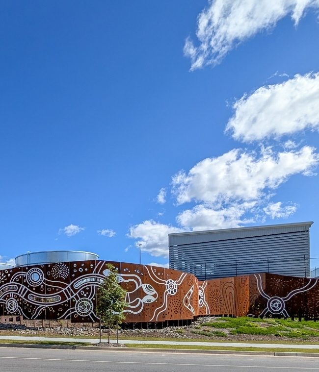 view of multiple screen panels on site demonstrating First Nations Artwork with beverage facility in background and surrounding landscapes showing greenery